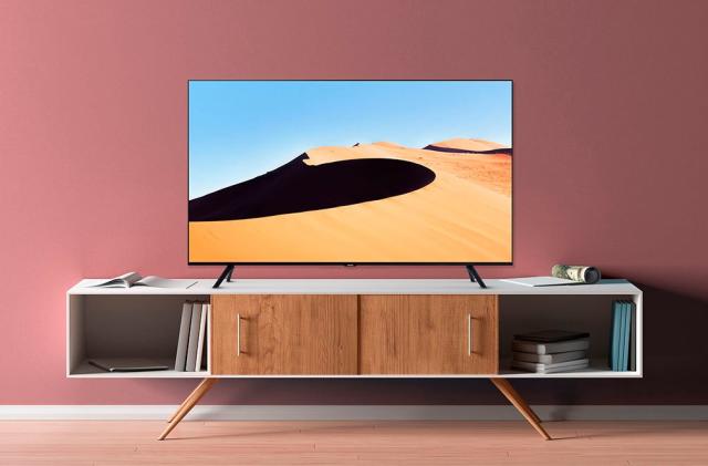 A press image of the Samsung TU690T 4K TV, displaying a TV on a white and brown cabinet against a light red wall. The image on the TV displays a desert dune.