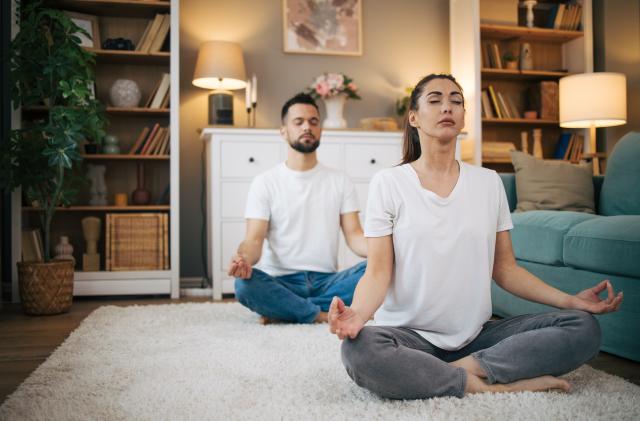 Picture of a man and a woman sitting on the floor in a modern apartment and practicing yoga. They wear white shirts.