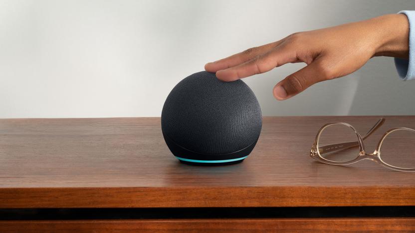 Amazon's Echo Dot speaker sits on a wooden nightstand. A person's hand is reaching out to touch the top of the speaker. Probably to snooze the alarm. 