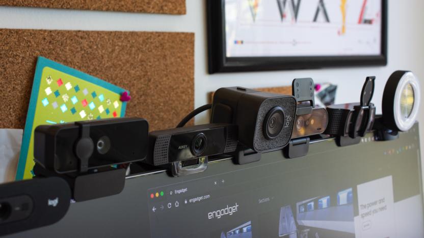 Seven webcams sitting on top of a computer monitor with cork boards on the wall behind them.
