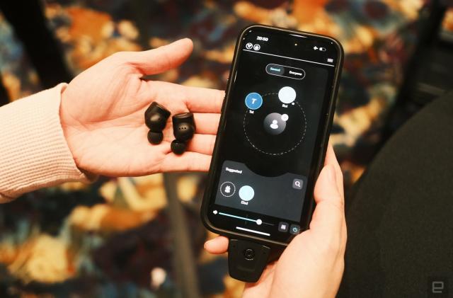 A pair of black earbuds on a person's palm, with their other hand holding up a phone with the OrCam Hear app on its screen.