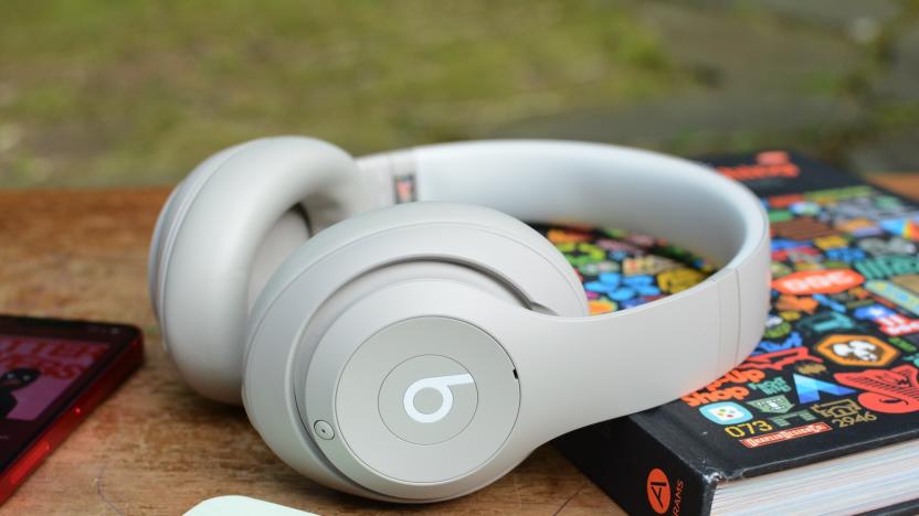 A white pair of Beats Studio Pro over-ear headphones sit on a wooden tabletop as it leans on a hardcover book.