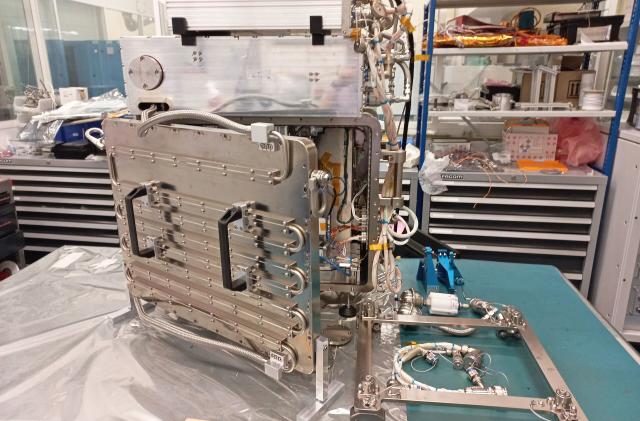 A raw metal 3D printer with lots of stainless steel and tubing that will be tested on the International Space Station sits on a cluttered workbench in a lab.