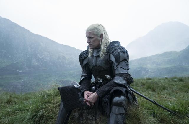 Still from House of the Dragon. A man with long white hair wears a suit of armor as he sits on a mound of grass. There are mountains in the background.