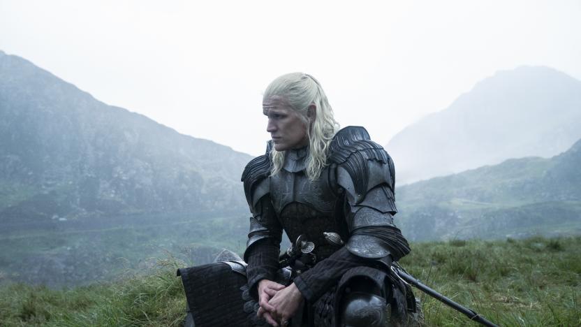 Still from House of the Dragon. A man with long white hair wears a suit of armor as he sits on a mound of grass. There are mountains in the background.