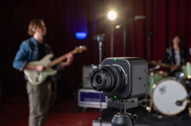 Lifestyle marketing image of the Logitech Mevo Core livestreaming camera. It sits on a tripod with a rock band playing in a studio behind them.