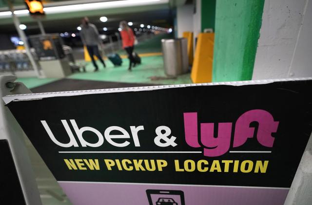 Passers-by pull luggage as they walk near a sign advising travelers of an Uber and Lyft pickup location at Logan International Airport, in Boston, Tuesday, Feb. 9, 2021. (AP Photo/Steven Senne)