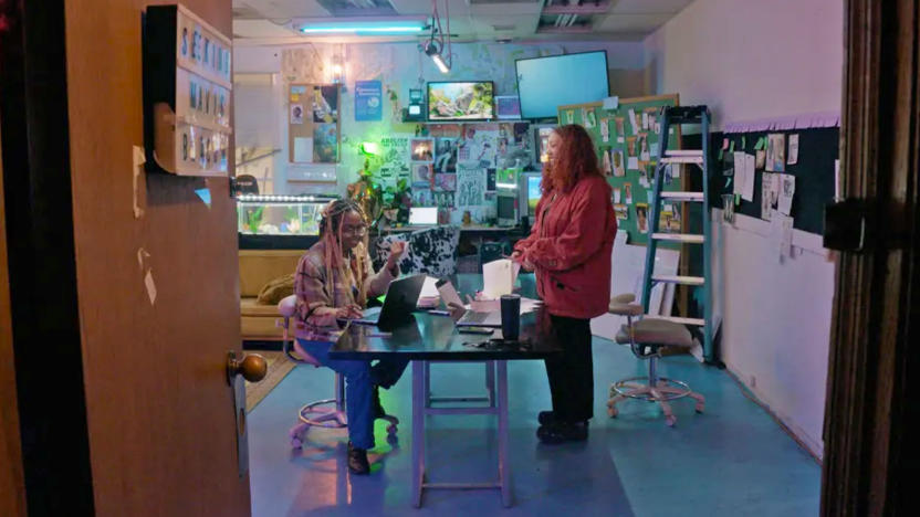 In this still from the documentary Seeking Mavis Beacon we see two women at a table, one sitting and one standing, surrounded by clutter from a ladder, to a fish tank.