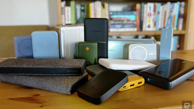Fourteen different power banks and battery packs from brands like Anker, Otterbox, and Mophie arranged on a wooden table with a bookcase in the background. 