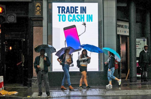 People with umbrellas on a city sidewalk are walking in front of an illuminated sign stating "Trade In to Cash In".