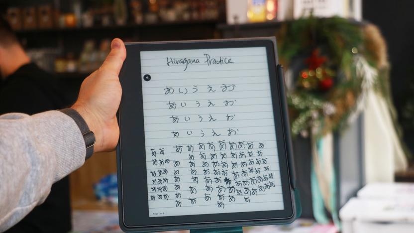The Amazon Kindle Scribe held up in mid-air by a hand gripping its right side. The screen shows the words "Hiragana Practice" at the top, followed by columns of Japanese script below.