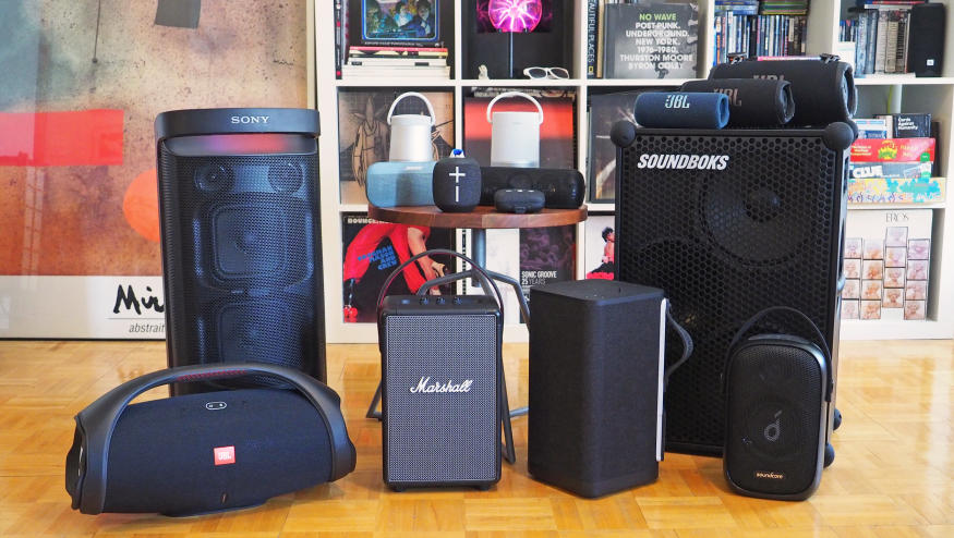 A group of portable Bluetooth speakers in front of a shelf full of books and stuff.