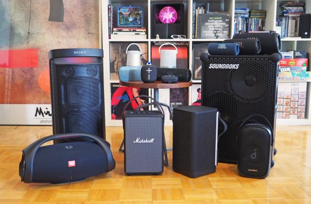 A group of portable Bluetooth speakers in front of a shelf full of books and stuff.