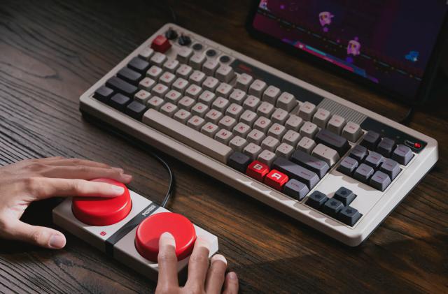 Product lifestyle photo of the 8BitDo Retro Mechanical Keyboard. The keyboard has Nintendo NES-inspired colors with deep (mechanical) keys. A pair of hands mash the two big red Super Buttons wired in front of it.