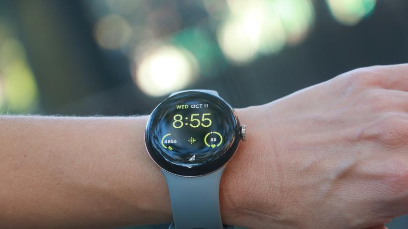 The Pixel Watch 2 on a person's wrist held in mid-air, with some blurred out greenery in the background.
