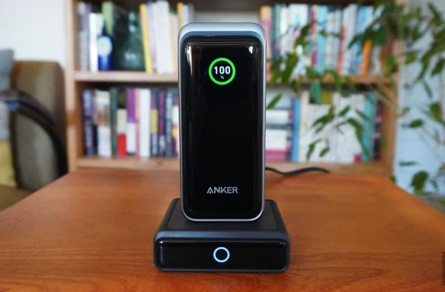 The Anker Prime power bank sits on its charging dock atop a wooden table. There are books and plants in the background. 
