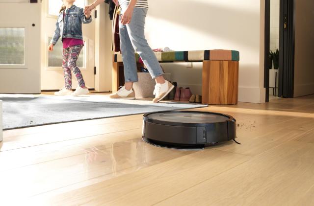 Marketing photo for the iRobot Roomba j5+. The robot vacuum rolls across a family's hardwood floor. In the background, a mother holds a daughter’s hand as they head out the front door.