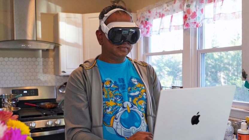 A man stands in front of his laptop in a kitchen while wearing the Apple Vision Pro AR/VR headset.
