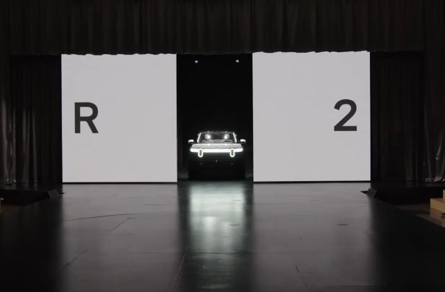 The Rivian R2 emerging from two withdrawing white panels on a stage at a launch event.