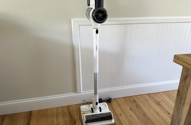 A white cordless stick vacuum sitting on its charging base. It's on wood tiled floor with light brown walls behind and the corner of a wooden table visible to the right.