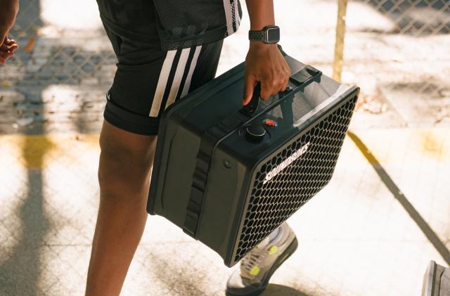 Someone in workout clothes carries a Soundboks Go Bluetooth speaker into a basketball court.