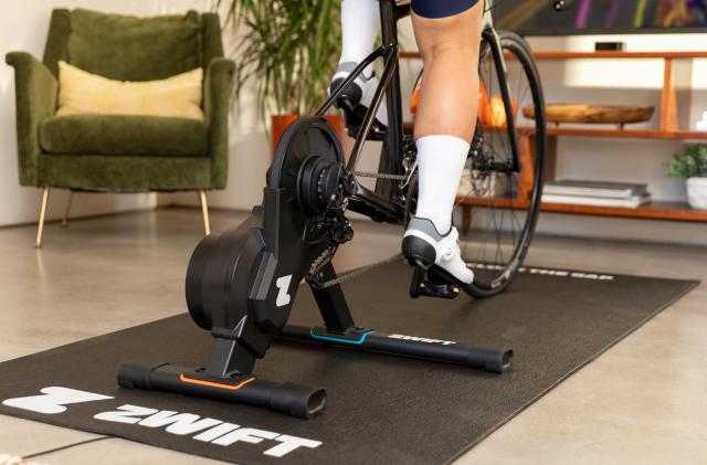 A Zwift Hub One bike trainer seen in use from behind. A person's bike is connected and indoors facing a TV with the Zwift app onscreen.