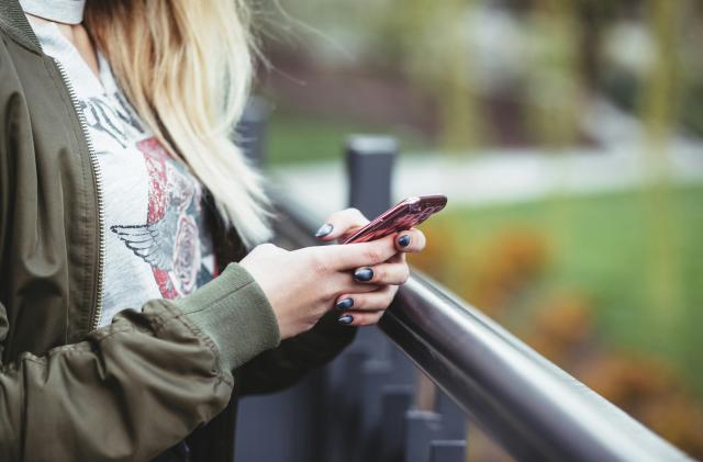 The torso of what appears to be a young woman holds a smartphone while standing next to a railing.