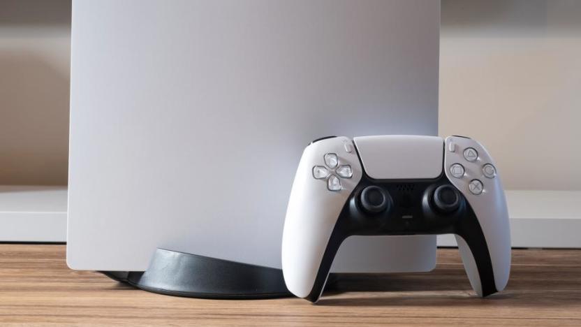 Closeup of a DualSense controller leaning against the PS5 console. Only the bottom part of the (upright) console is visible. Wood-grained desk with blurred background.