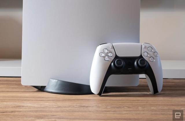 Closeup of a DualSense controller leaning against the PS5 console. Only the bottom part of the (upright) console is visible. Wood-grained desk with blurred background.