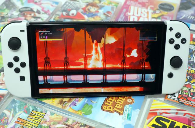 Photo of the Nintendo Switch OLED sitting on a colorful array of game cartridge cases. Its screen shows a fiery scene.