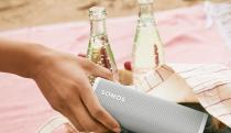 A closeup of a person's hand taking a Sonos Roam speaker out of a bag with bottles of a carbonated beverage behind it