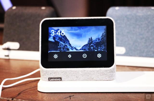 A gray Lenovo Smart Clock 2 on a wireless charging dock with its screen facing the camera. A row of options for volume, brightness, do not disturb and settings take up the bottom of the display.  In the background are a black and a blue Smart Clock 2 on the left and right respectively.