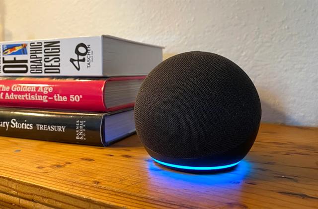 The Amazon Echo Dot rests on a brown woodside table next to a stack of three books, with the status indicator light around the bottom of the device glowing blue.