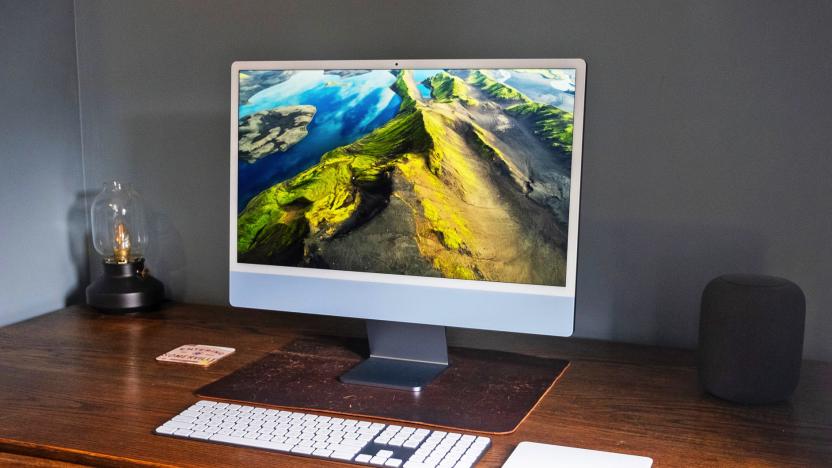 The 2023 Apple iMac with a 24-inch screen and the M3 chip in ice blue sits on a worn wooden corner desk next to a small lamp and an Apple HomePod.