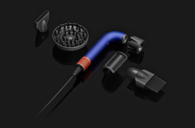 The components of the Dyson Supersonic R hairdryer are laid out next to each other on a black surface. in the middle is the main J-shaped tubular dryer body with its several attachments.